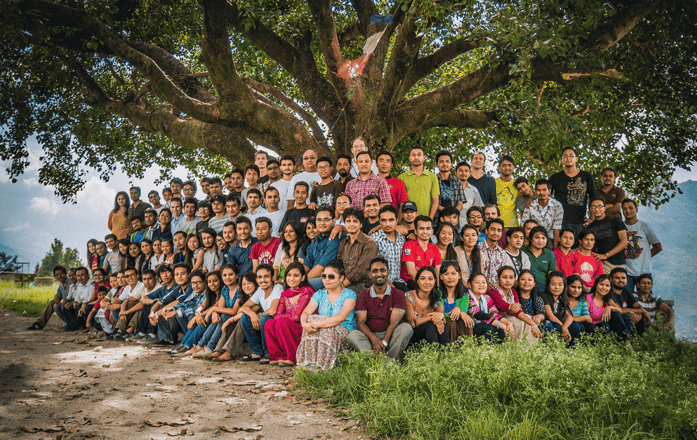 A group of approximately 100 CloudFactory core team members in Nepal sits under a large, shady tree for a photoshoot.